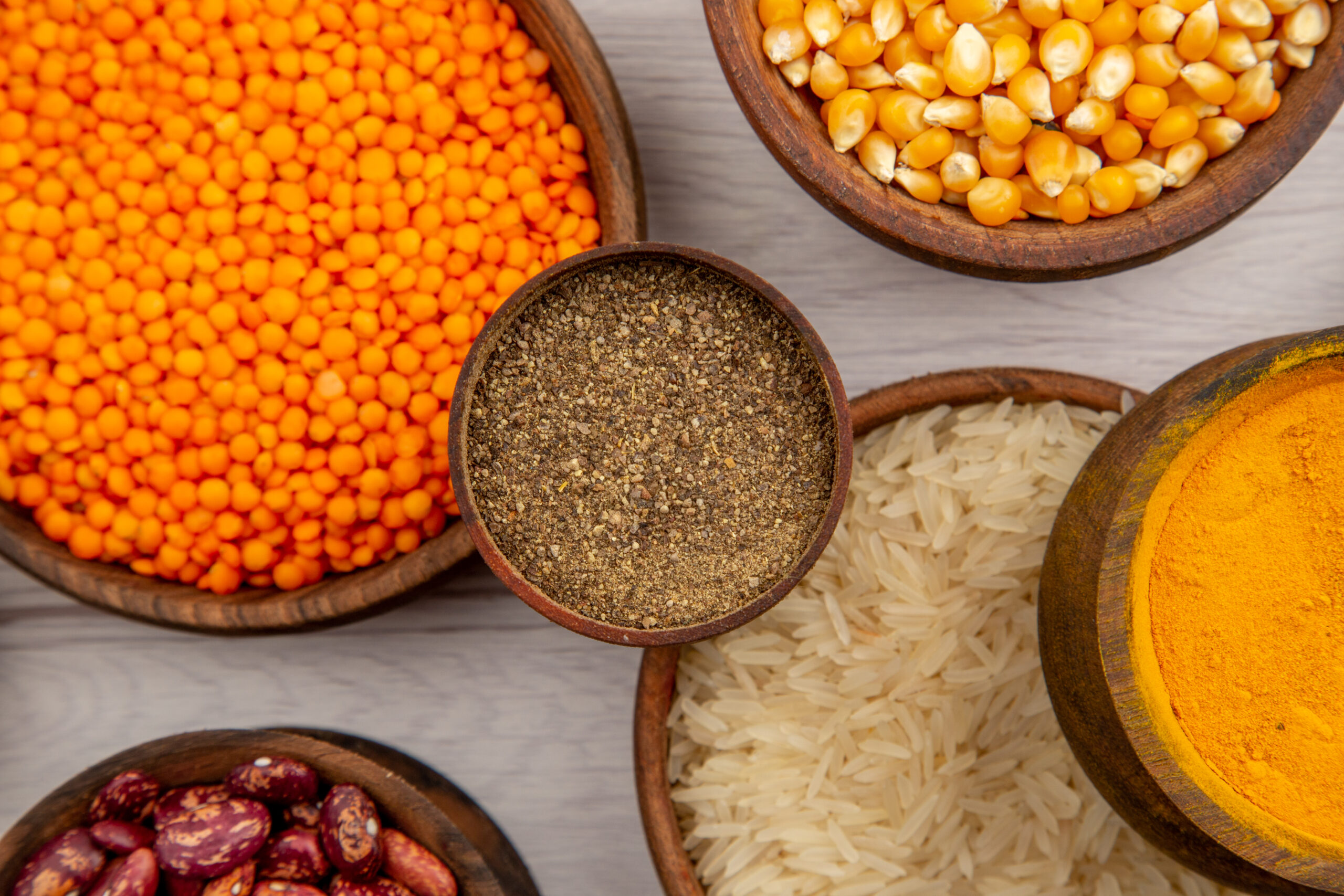 top-view-wooden-bowls-with-rice-pumpkin-seeds-black-pepper-powder-turmeric-lentiles-grey-background